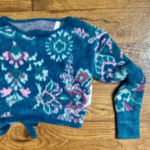 May include: A teal blue fuzzy sweater with a pink, green, and white floral pattern. The sweater has a tie at the bottom.