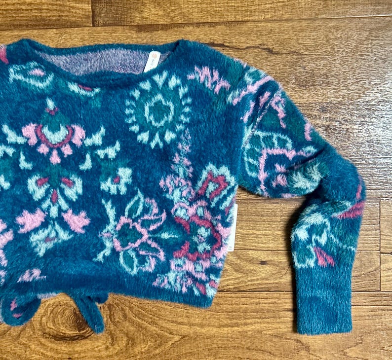 May include: A teal blue fuzzy sweater with a pink, green, and white floral pattern. The sweater has a tie at the bottom.