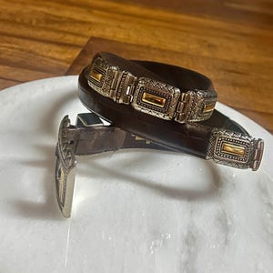 May include: A brown leather belt with silver and gold-toned rectangular buckle and accents. The belt features a series of decorative silver and gold-toned rectangular links. The buckle and links have an ornate, textured design.