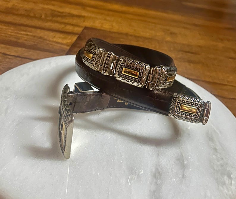 May include: A brown leather belt with silver and gold-toned rectangular buckle and accents. The belt features a series of decorative silver and gold-toned rectangular links. The buckle and links have an ornate, textured design.