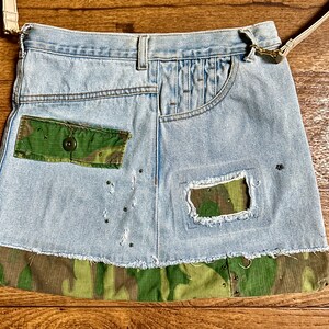 May include: A light blue denim bag with a camouflage fabric trim. The bag has a flap with a pocket and a large pocket on the front. The bag has a brown leather strap.