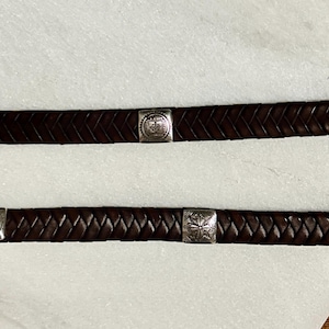 K&ouml;nnte beinhalten: Zwei dunkelbraune Lederg&uuml;rtel mit geflochtenem Design. Jeder G&uuml;rtel hat quadratische, silberfarbene Metallakzente mit dekorativen Mustern. Die G&uuml;rtel werden auf einer wei&szlig;en Oberfl&auml;che mit Holzgrund gezeigt.