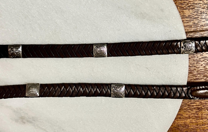 K&ouml;nnte beinhalten: Zwei dunkelbraune Lederg&uuml;rtel mit geflochtenem Design. Jeder G&uuml;rtel hat quadratische, silberfarbene Metallakzente mit dekorativen Mustern. Die G&uuml;rtel werden auf einer wei&szlig;en Oberfl&auml;che mit Holzgrund gezeigt.