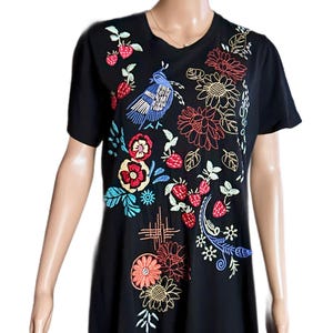 Puede incluir: Un vestido tipo camiseta negro de manga corta con bordados coloridos. El dise&ntilde;o incluye un p&aacute;jaro azul, fresas rojas y varios motivos florales en tonos naranja, amarillo, rojo y azul, distribuidos por toda la parte frontal de la prenda.