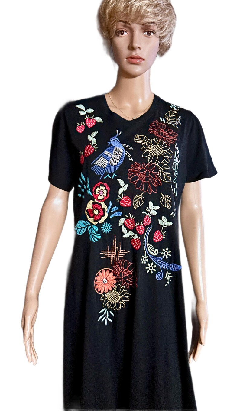 Puede incluir: Un vestido tipo camiseta negro de manga corta con bordados coloridos. El dise&ntilde;o incluye un p&aacute;jaro azul, fresas rojas y varios motivos florales en tonos naranja, amarillo, rojo y azul, distribuidos por toda la parte frontal de la prenda.