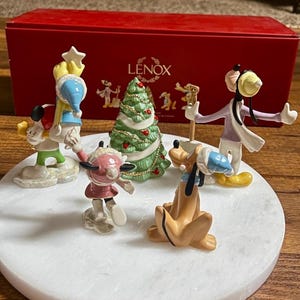 Może przedstawiać: Kolekcja figurek świątecznych Disneya od Lenoxa, w tym Myszka Miki, Myszka Minnie, Goofy i Pluto, ustawionych wok&oacute;ł udekorowanej choinki. Figurki znajdują się na białej powierzchni przypominającej marmur, z czerwoną skrzynką Lenox w tle.