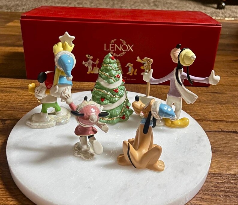 Może przedstawiać: Kolekcja figurek świątecznych Disneya od Lenoxa, w tym Myszka Miki, Myszka Minnie, Goofy i Pluto, ustawionych wok&oacute;ł udekorowanej choinki. Figurki znajdują się na białej powierzchni przypominającej marmur, z czerwoną skrzynką Lenox w tle.