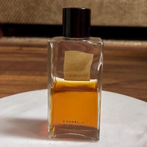 May include: A rectangular perfume bottle with a black cap. The clear glass bottle contains a golden-brown liquid. The label is a light tan color with text. The bottom of the bottle reads "CHANEL Inc."