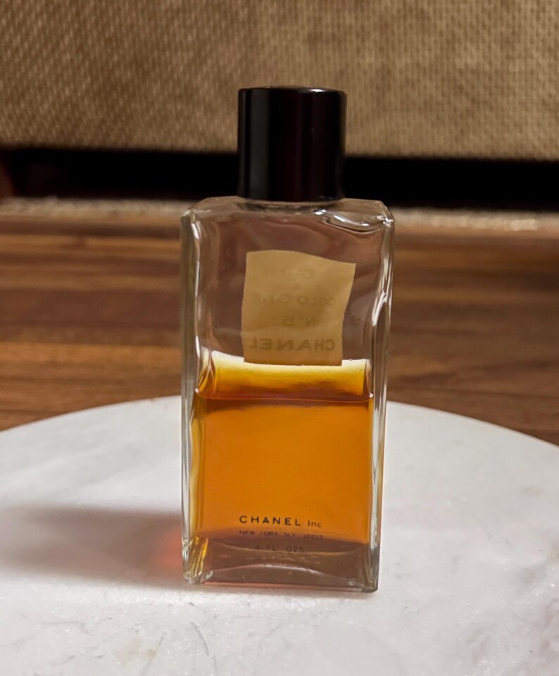 May include: A rectangular perfume bottle with a black cap. The clear glass bottle contains a golden-brown liquid. The label is a light tan color with text. The bottom of the bottle reads "CHANEL Inc."