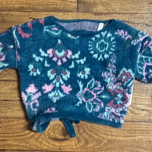 May include: A teal blue fuzzy sweater with a pink, green, and white floral pattern. The sweater has a round neckline and long sleeves.