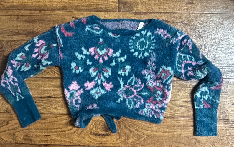 May include: A teal blue fuzzy sweater with a pink, green, and white floral pattern. The sweater has a round neckline and long sleeves.