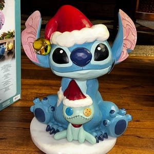 May include: A blue Stitch figurine wearing a red Santa hat with white trim and a gold bell. A smaller Stitch figure with a stitched face and a red hat is in front. The figurine sits on a white base.
