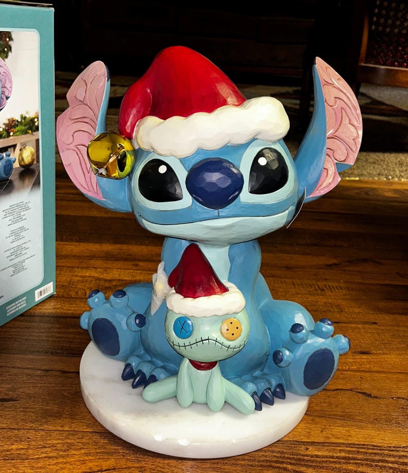 May include: A blue Stitch figurine wearing a red Santa hat with white trim and a gold bell. A smaller Stitch figure with a stitched face and a red hat is in front. The figurine sits on a white base.
