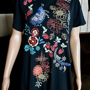 Puede incluir: Una camiseta negra de manga corta con bordados coloridos de un p&aacute;jaro azul, fresas rojas y varias flores en tonos amarillos, rojos y azules. El dise&ntilde;o est&aacute; distribuido por la parte delantera de la prenda, mostrando un detallado trabajo de costura.
