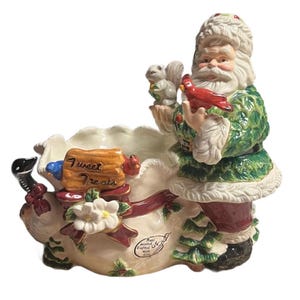 May include: A ceramic Santa Claus figurine with a white beard and fur-trimmed hat and coat. He holds a red cardinal and a squirrel. The base is a white bag with a red ribbon, a sign that reads "Sweet Treats", and a white flower.