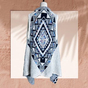 May include: A sleeveless knit vest featuring a large, central diamond-shaped pattern on the back. The design incorporates black, white, and iridescent blue and purple geometric shapes. The vest has a textured, off-white base fabric throughout.