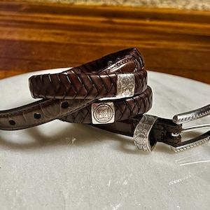 K&ouml;nnte beinhalten: Ein Stapel aus drei braunen Lederg&uuml;rteln mit silbernen Schnallen und Akzenten. Die G&uuml;rtel sind geflochten und haben dekorative silberne Verschl&uuml;sse. Die Schnalle ist silberfarben mit einem verzierten Design.