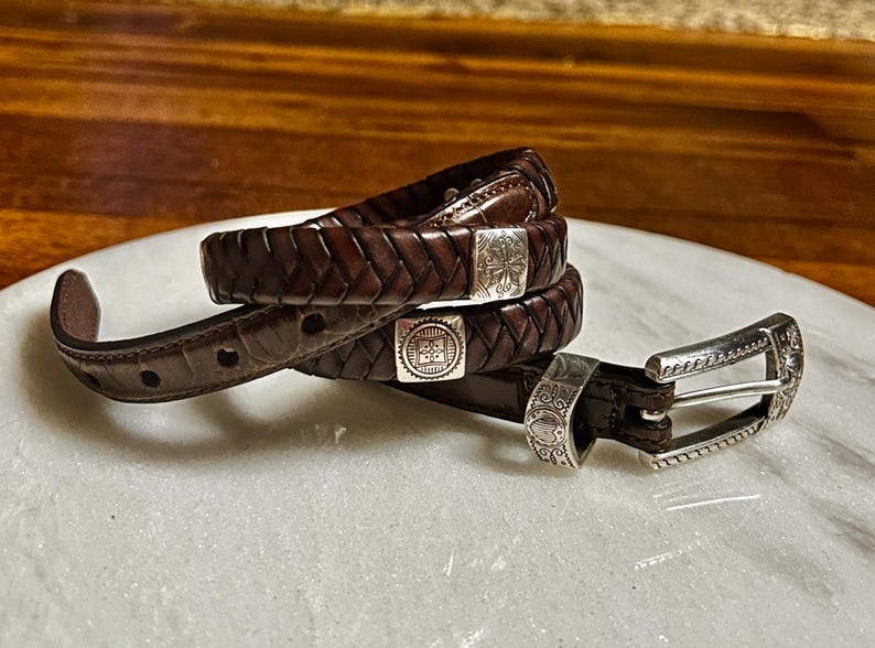 K&ouml;nnte beinhalten: Ein Stapel aus drei braunen Lederg&uuml;rteln mit silbernen Schnallen und Akzenten. Die G&uuml;rtel sind geflochten und haben dekorative silberne Verschl&uuml;sse. Die Schnalle ist silberfarben mit einem verzierten Design.