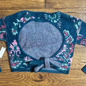 May include: A teal blue fuzzy sweater with a pink and green floral pattern. The sweater has a large open back with a tie detail.