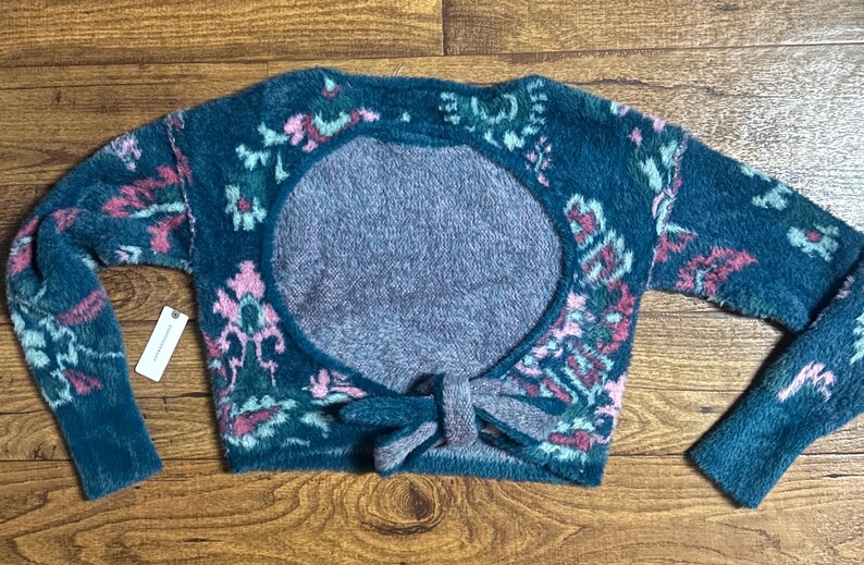 May include: A teal blue fuzzy sweater with a pink and green floral pattern. The sweater has a large open back with a tie detail.