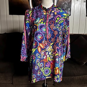 May include: A long-sleeved tunic with a vibrant, colorful paisley pattern. The tunic features a mandarin collar, a partial button placket, and pink stripes down the sleeves. The design incorporates shades of blue, purple, yellow, and green.