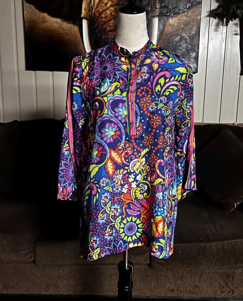 May include: A long-sleeved tunic with a vibrant, colorful paisley pattern. The tunic features a mandarin collar, a partial button placket, and pink stripes down the sleeves. The design incorporates shades of blue, purple, yellow, and green.