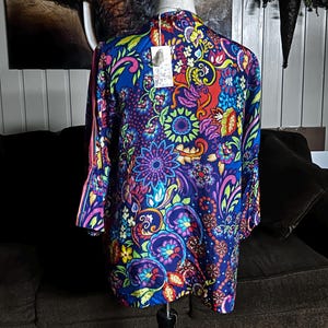 May include: A long-sleeved, vibrant blue top with a colorful floral pattern in shades of purple, red, yellow, and green. The top is displayed on a mannequin, showcasing its design and fit. The sleeves have a flared design.