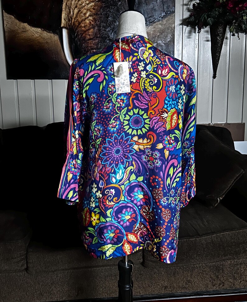 May include: A long-sleeved, vibrant blue top with a colorful floral pattern in shades of purple, red, yellow, and green. The top is displayed on a mannequin, showcasing its design and fit. The sleeves have a flared design.