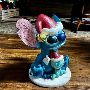 May include: A blue and turquoise Stitch figurine wearing a Santa hat and holding a smaller Stitch figure. The larger Stitch has a gold bell on its ear and a pink wing. The figurine is on a white circular base.
