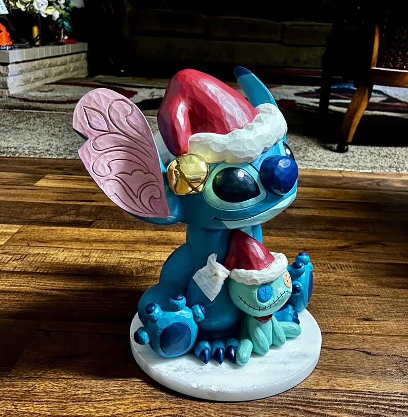May include: A blue and turquoise Stitch figurine wearing a Santa hat and holding a smaller Stitch figure. The larger Stitch has a gold bell on its ear and a pink wing. The figurine is on a white circular base.