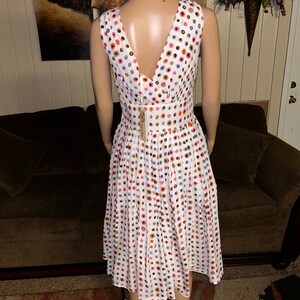 May include: A sleeveless white dress with a V-neck and a pleated skirt. The dress is adorned with colorful polka dots in shades of blue, pink, orange, and brown. A tag is attached to the dress. The dress is displayed on a mannequin.