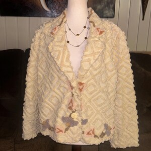 May include: A cream-colored, fuzzy jacket with a geometric pattern and floral embroidery. The jacket has a button closure and long sleeves.