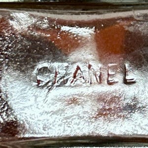 May include: Close-up of a clear glass object with the embossed word "CHANEL". The glass has a textured surface and a slight amber hue, with light reflections creating highlights and shadows.