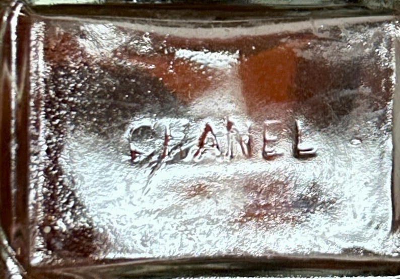 May include: Close-up of a clear glass object with the embossed word "CHANEL". The glass has a textured surface and a slight amber hue, with light reflections creating highlights and shadows.