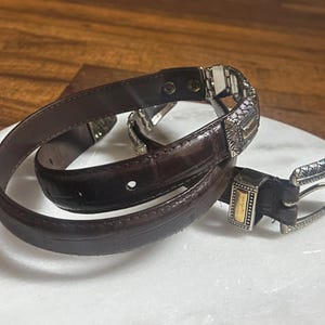 May include: A dark brown leather belt with a silver and gold buckle. The belt is coiled on a white surface, showcasing its intricate design and texture. The buckle features detailed patterns and a contrasting gold accent.