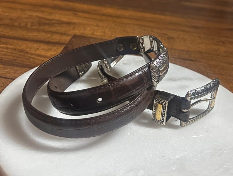May include: A dark brown leather belt with a silver and gold buckle. The belt is coiled on a white surface, showcasing its intricate design and texture. The buckle features detailed patterns and a contrasting gold accent.