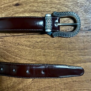 Puede incluir: Un cintur&oacute;n de cuero marr&oacute;n con una hebilla plateada con un dise&ntilde;o intrincado. La hebilla es adornada y tiene un patr&oacute;n detallado.