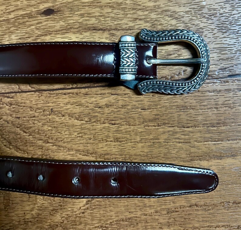 Puede incluir: Un cintur&oacute;n de cuero marr&oacute;n con una hebilla plateada con un dise&ntilde;o intrincado. La hebilla es adornada y tiene un patr&oacute;n detallado.