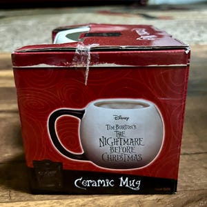 Puede incluir: Una taza de cer&aacute;mica en una caja roja con el texto "Tim Burton's The Nightmare Before Christmas". La taza es blanca con un asa negra. La caja tiene un dise&ntilde;o de remolino.