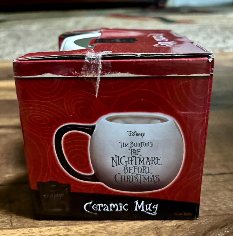 Puede incluir: Una taza de cer&aacute;mica en una caja roja con el texto "Tim Burton's The Nightmare Before Christmas". La taza es blanca con un asa negra. La caja tiene un dise&ntilde;o de remolino.