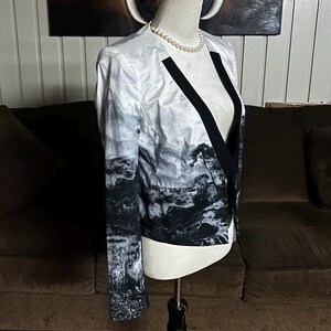 May include: A white and black landscape print blazer with black lapels and a black trim. The blazer is displayed on a mannequin with a pearl necklace. The jacket has long sleeves and a tailored fit.
