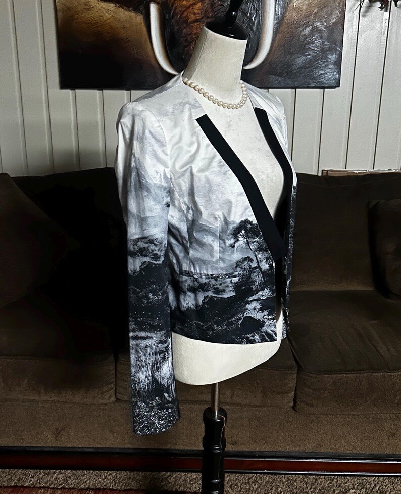 May include: A white and black landscape print blazer with black lapels and a black trim. The blazer is displayed on a mannequin with a pearl necklace. The jacket has long sleeves and a tailored fit.
