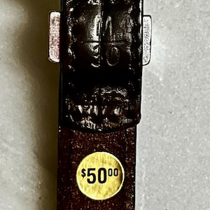 K&ouml;nnte beinhalten: Nahaufnahme eines braunen Lederg&uuml;rtels mit einer silbernen Schnalle. Der G&uuml;rtel hat eine strukturierte Oberfl&auml;che und einen goldenen Aufkleber mit dem Text "$5000". Der G&uuml;rtel ist ein klassisches Accessoire.