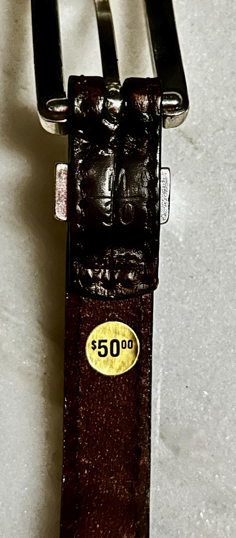 K&ouml;nnte beinhalten: Nahaufnahme eines braunen Lederg&uuml;rtels mit einer silbernen Schnalle. Der G&uuml;rtel hat eine strukturierte Oberfl&auml;che und einen goldenen Aufkleber mit dem Text "$5000". Der G&uuml;rtel ist ein klassisches Accessoire.