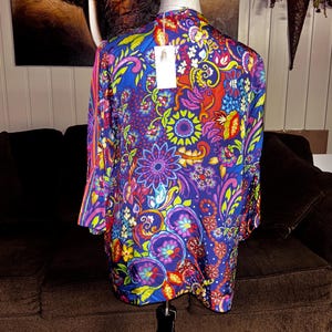 May include: A vibrant, long-sleeved top with a colorful floral pattern in shades of blue, purple, red, and yellow. The garment is displayed on a mannequin, showcasing its design. A tag hangs from the neckline.