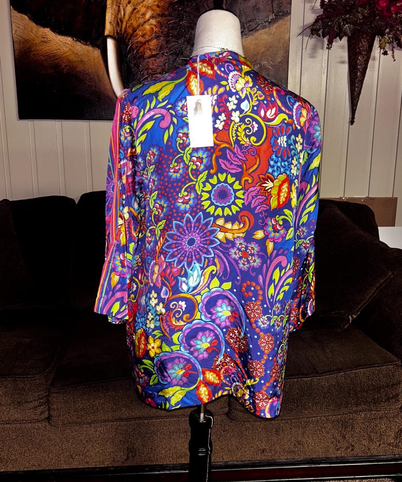 May include: A vibrant, long-sleeved top with a colorful floral pattern in shades of blue, purple, red, and yellow. The garment is displayed on a mannequin, showcasing its design. A tag hangs from the neckline.