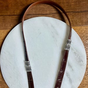 May include: A brown leather purse strap with silver-toned metal accents. The strap is made of a dark brown, textured leather and has decorative metal bands near the ends. The background is a white marble surface and a wooden surface.