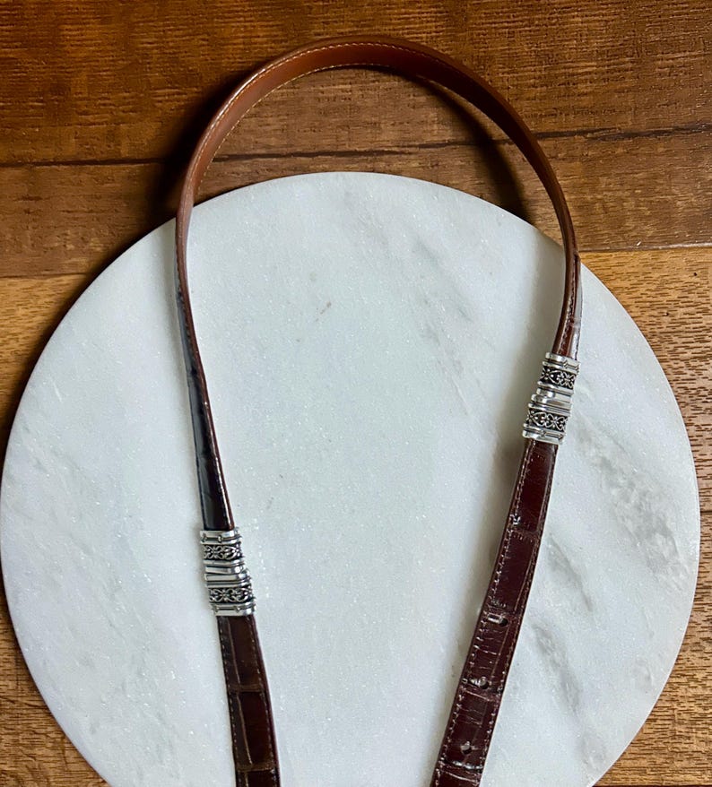 May include: A brown leather purse strap with silver-toned metal accents. The strap is made of a dark brown, textured leather and has decorative metal bands near the ends. The background is a white marble surface and a wooden surface.