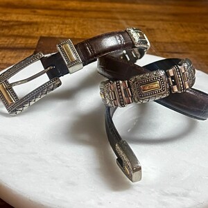 May include: Two brown leather belts with ornate silver and gold-toned buckles. The belts are displayed on a white marble surface, showcasing their intricate designs and textures. The buckles feature detailed patterns.
