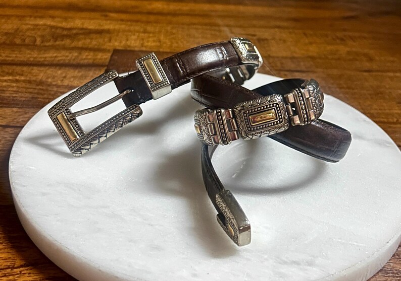 May include: Two brown leather belts with ornate silver and gold-toned buckles. The belts are displayed on a white marble surface, showcasing their intricate designs and textures. The buckles feature detailed patterns.